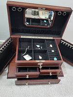 Open jewelry box showing multiple compartments with assorted jewelry pieces like earrings, pendants, and necklaces on black velvet lining, upper lid has a mirror and ring hangers.