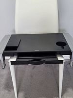 Black Fellowes monitor riser with adjustable chrome legs, two pen trays on left side, round cup holder on right, and pull-out drawer underneath.