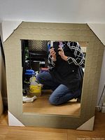 Full view of square mirror with beige textured frame and corner protectors, showing size relative to person kneeling.