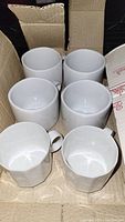 Six white porcelain cups arranged in two rows, showing their standard size, shape, and handle design inside a cardboard box.