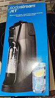 Front view of SodaStream Jet soda maker on its box showing the model and branding with a glass of sparkling water.