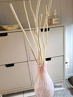 Full view of tall pink glass vase with white wood branches inside, set against a white cabinet and beige carpet.