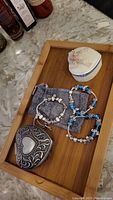 Photo showing two heart-shaped trinket boxes (one large silver-tone, one smaller white and blue) and three costume jewelry charm bracelets displayed on a wooden tray.