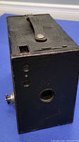 Front view of the black box camera showing lens, viewfinder, and leather strap on top.