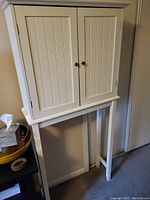 Front view of white over toilet bathroom cabinet showing two beadboard panel doors with silver knobs and elevated legs.
