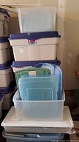 Stack of plastic storage bins and many lids inside a clear bin on top of gray and white bins in garage setting.