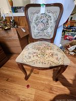 Front view of chair showing needlepoint upholstery with classical scene on backrest and floral pattern on seat, wood frame with carving.