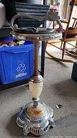 Full view of chrome-plated floor ashtray on concrete floor, showing scalloped metal base, cream marble mid-section and slender metal column supporting top tray with black ashtray insert and lighter handle.