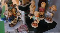 Group photo showing six Goebel Hummel figurines arranged on black tiered stand and one on glass shelf. The children are dressed in winter attire and some hold musical instruments or toys.