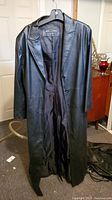 Black leather ladies trench coat hanging on a hanger, showing the full length and design.