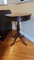 Angled side view of wooden round side table showing turned pedestal legs and drawer with metal pull.