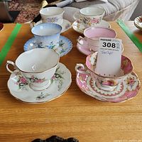Six bone china tea cups with matching saucers displayed on wooden surface, various floral and gold designs visible.