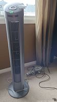 Full view of Seville Classics tower fan standing on carpeted floor next to window and curtain, with extension cords coiled beside base.