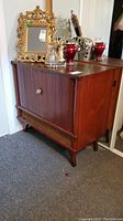 Front-left angle of vintage wooden stereo cabinet with roll-away tambour doors and tapered legs. Top has decorative items (not included).