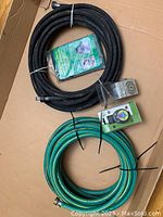 Lot contents on cardboard: black soaker hose coiled with two metal fittings, green garden hose coiled with metal fittings, two garden water timers in packaging, green garden apron, oscillating sprinkler with metal body and black base.