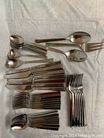 Sets of stainless steel forks, knives, tablespoons, and teaspoons alongside serving fork, spoon, and slotted spoon on a white fabric surface.