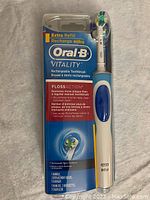 Photo of Oral-B Vitality Floss Action electric toothbrush in original sealed packaging showing handle, brush heads, charger, and branding information.
