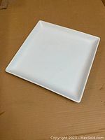 White square ceramic platter placed on a brown surface, showing the front and angled side view to highlight shape and size.