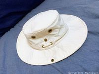 Tilley off white outdoor hat resting on blue fabric showing brim and crown with metal eyelets and chin snaps.