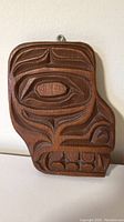 Front view of a wood carving depicting an Indigenous styled bear face with detailed geometric features and natural wood finish.