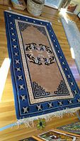 Full view of rectangular area rug with beige center and blue border featuring geometric and symbolic patterns with fringed edges on hardwood floor.