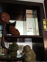 Shelf segment showing red goldstone Buddha head bust, brown carved piece, jadeite seated Buddha and cream-colored Budai figurine