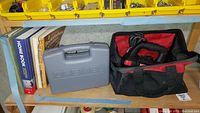 Wide view of shelf containing books about home maintenance, carpentry, and composting, Dremel kit in gray case, and soft carrying bag with Craftsman cutting tool.