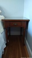 Side view of wooden end table with visible drawer, turned legs, and glass top next to a bed.