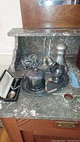 Silver plated sugar bowl, creamer, oval tray, canister, shaker, two hand bells, and a small pitcher arranged on marble countertop.
