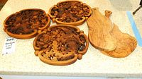 Five wooden trivets on a countertop including three with detailed handcarvings and two smooth wooden cutting or serving boards.