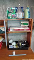 Wooden shelf containing various spray bottles (Windex, garden products), first aid kit box, Bosch paper cutter packaging, and other miscellaneous tools and materials.
