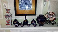 Full view of all items on shelf including sake set with crane motifs, Imari-style rectangular vase, large blue jar, and decorative plate on stand.