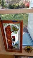 Two stained glass panels, each featuring a woodpecker on a tree trunk with leaves, shown leaning on window with sunlight behind.