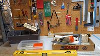 Workshop table with assorted tools including spirit level, hand saws, boxes with hardware and tools, clamps, pliers, and pegboard with hanging tools.