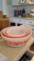 Stacked three Pyrex mixing bowls on counter showing orange and pink bird and flower design on largest and smallest bowl, solid orange middle bowl
