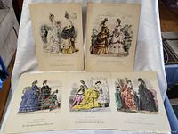 Grouped view of five antique 1800s fashion dressmaking prints with women in elaborate dresses in various colors and outdoor scenes.