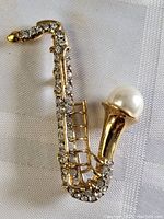 Full view of the gold-tone saxophone brooch pin with clear rhinestones and large white faux pearl at the bell end.