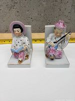 Pair of porcelain bookends with Asian figurine designs; one reading a book and one playing an instrument, shown with ruler for scale.