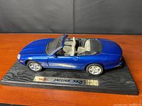 Front-side view of blue Jaguar XK8 model car on wooden base showing light interior and wheels.