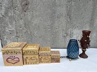 Four sizes of square decorative wooden boxes with various motifs and text plus a blue textured glass vase and a brown candle holder with beads.