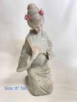 Porcelain geisha figurine holding a fan, detailed floral kimono, styled hair with pink flowers, 8 inches tall.