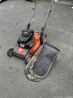 Side view of the Husqvarna self-propelled lawn mower with fabric bagger placed beside it on asphalt.