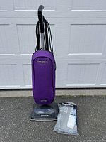 Front view of purple Oreck upright vacuum cleaner with extra filter bag placed nearby on ground.