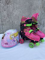 Pair of adjustable pink and green Roller Derby roller skates alongside a children's helmet with Disney Frozen characters Elsa and Anna on it, photographed against a rough gray concrete wall.