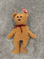 Photo of brown TY Beanie Babies teddy bear plush toy lying on grey carpet, showing full body with original tag on ear and red ribbon around neck.
