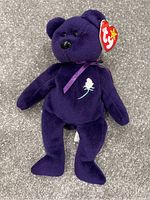 Full view of the purple Beanie Baby Princess bear with its Ty tag visible, lying on a gray textured surface.