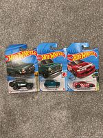 Photo of three new Hot Wheels cars in original packaging, showing black off-road vehicle, teal-green compact car, and red sports car with decals.