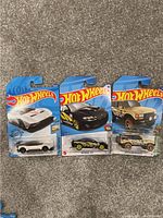 Photo shows three Hot Wheels die-cast cars still in their original sealed packaging against a carpeted background.