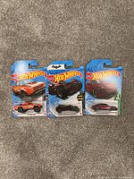 Three Hot Wheels toy cars in original sealed packaging. From left to right: orange muscle car, black Batman-themed vehicle, and red sports car.