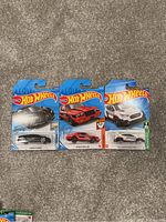 Three Hot Wheels toy cars still in their original packaging, featuring Chevrolet Camaro, Firebird OE, and Porsche Cayenne Turbo models.